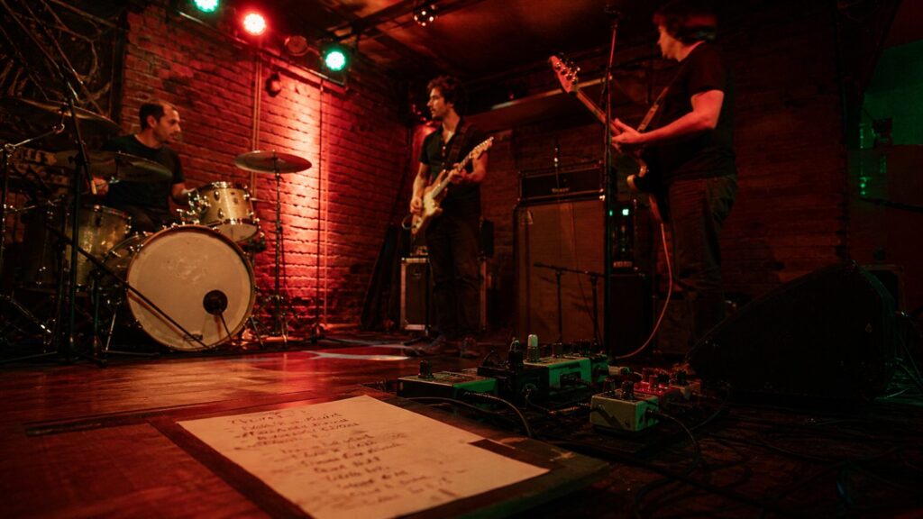 Lista de canciones manuscrita junto a una guitarra, pedales y luces rojas y verdes de ensayo